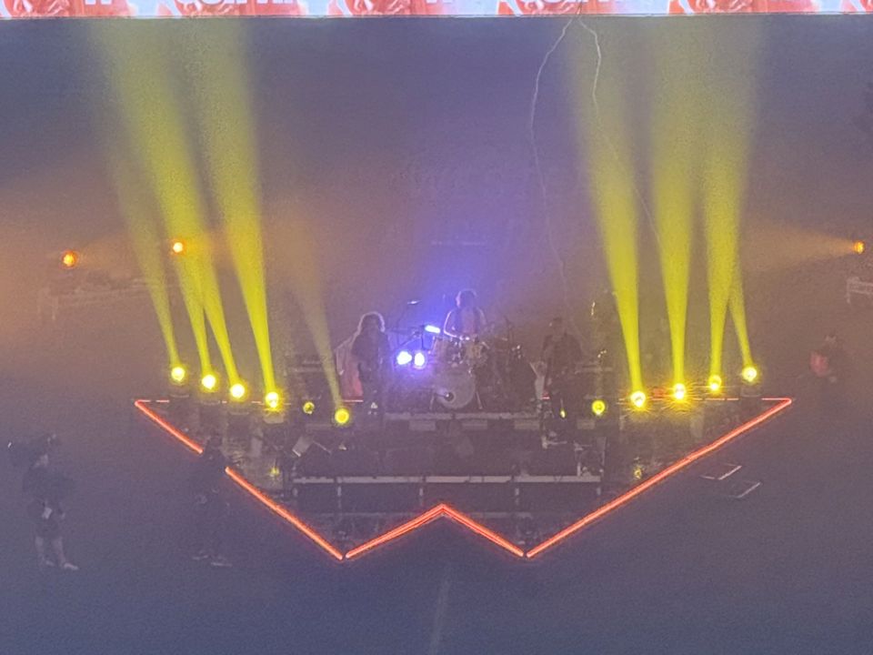 Triangle stage from CorpraHire's Wolfmother stage project, outlined in orange LED lighting, with a band performing under yellow spotlight beams in a darkened stadium