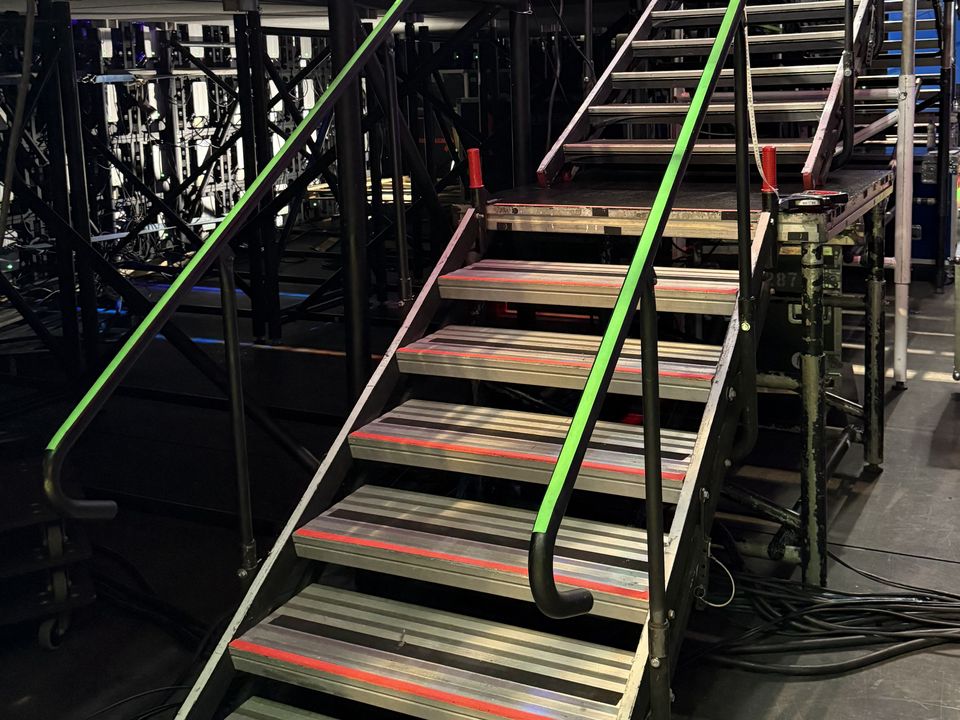 Two flights of aluminium stage stairs with green handrails and red-edged treads rising to a high stage deck in an arena