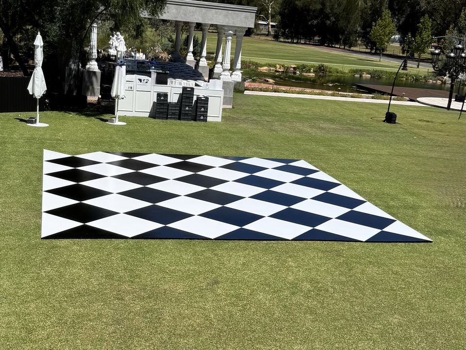 Black and white diamond-pattern dance floor laid on a lawn at a garden venue, with white columns, trees and a pond behind
